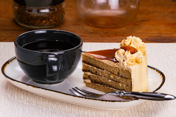 Side view of a cup of black coffee and a slice of coffee layered cake garnished with cream and almond in white ceramic tray with dark edge and a metal fork on wooden table.