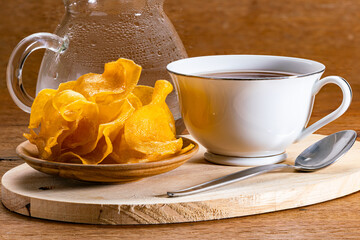 Closeup view of sweet crispy potato chips in wooden plate with a white ceramic cup of hot tea and a metal spoon on wooden cutting board and a transparent glass pitcher on wooden table.