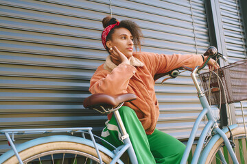 Outdoor fashion portrait of pretty african woman sitting on bike, leaning on the wall