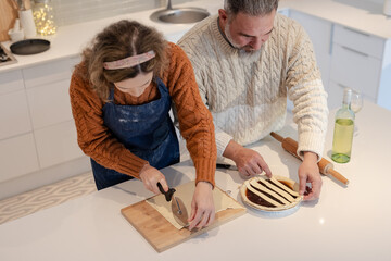 Couple rolling pastry dough with rolling pin, placing lattice strips on fruit pie at kitchen island