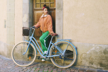 Outdoor fashion portrait of pretty african woman sitting on bike, leaning on the wall