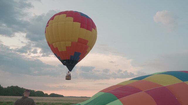 crew prepping second hot air balloon while earlier colorful balloon hovers above grassy open field at sunset during preflight setup capturing teamwork and scenic adventure mood under dramatic sky