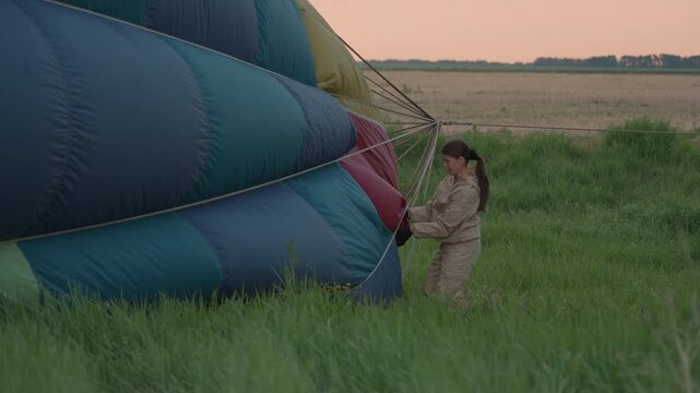 woman working on hot air balloon envelope in tall grass preparing envelope for inflation during sunset field preflight operation showcasing focus teamwork and vibrant fabric