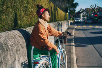 Outdoor fashion portrait of pretty african woman sitting on bike, leaning on the wall