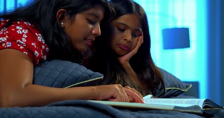 Indian sibling sisters reading book while lying on bed enjoying quiet story time together. Asian little girls wearing colorful clothes focused on book indoors in cozy bedroom during leisure moments - Powered by Adobe