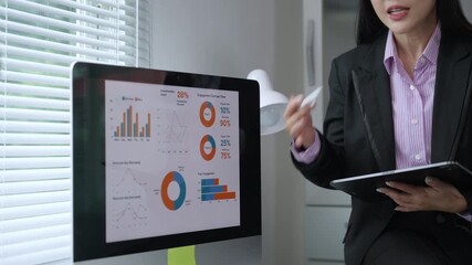 Professional businesswoman analyzing financial data, presenting charts and graphs using digital pen while holding tablet, working at modern office workspace with computer monitor - Powered by Adobe