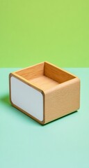 A small, light wood box with a white panel stands on a split green background. The box is empty and rectangular with rounded corners. The overall scene is clean and minimalist