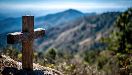 Wooden cross stands on a rocky hilltop, overlooking blurred rolling mountains under a clear blue sky. Tranquil, simple, and peaceful scene