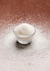  A small glass bowl overflows with sparkling white granulated sugar, surrounded by scattered sugar crystals on a warm wooden surface