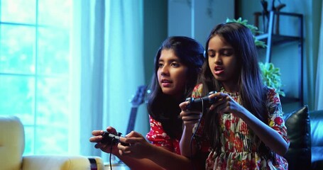 Indian little sisters playing video game together while sitting on sofa at home, Asian sibling girls enjoying gaming with joystick, cheerful childhood fun, electronic entertainment, family bonding - Powered by Adobe