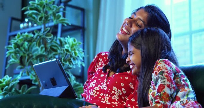 Indian little sisters laughing watching funny video on tablet computer while sitting on sofa at home, Asian sibling girls enjoying digital entertainment, childhood fun, happiness, family bonding