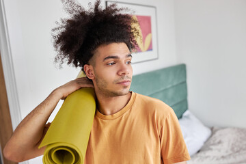 Young attractive dark-skinned guy of 25 in orange home t-shirt holding rolled yellow mat over his shoulder leaving his bedroom to train outdoor in summer morning. Relaxation, home fitness concept