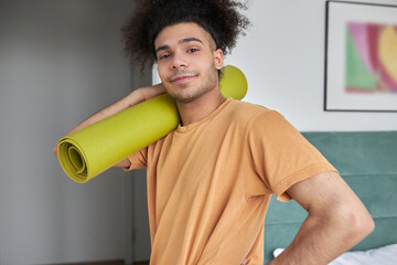 Athletic black photogenic man in orange t-shirt carrying rolled yellow mat over his shoulder after having intensive home workout in his bedroom after waking up in morning, before going to work
