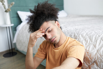 Horizontal image of afro guy sitting on floor in his bedroom and feeling unwell, holding his...