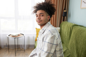 Stylish attractive young boy of 20s sitting on sofa in his girlfriend's living-room and looking at camera waiting for her getting ready to go for date to watch cinema or to walk outside