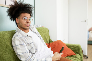 Handsome young guy of black ethnicity with stylish haircut having rest sitting on sofa with smartphone in hands, chatting with his friends and making appointment to meet in downtown