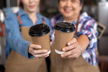 Two women wearing aprons hold takeaway coffee cups toward the camera with warm smiles, symbolizing...