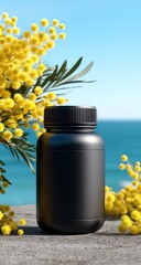 A black cylindrical bottle stands outdoors near water, framed by bright yellow flowers, sitting on a neutral stone surface under a clear sky