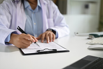 Close up of female doctor filling out medical form on clipboard at desk