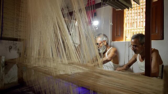 Mid black shot of Indian artisans weaving a Banarasi saree with intricate gold thread work on a traditional handloom in Varanasi, India. Shot shows detailed weaving process and craftsmanship