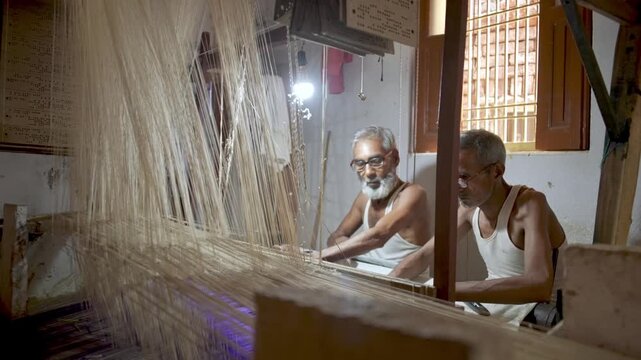Profile view of two artisans weaving Banarasi silk fabric on a handloom in Varanasi, India. Traditional textile craftsmanship with fine threads and patterns in a detailed indoor setting.
