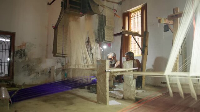 Wide angle view of two men weaving silk fabric using handloom machines inside a workshop in Varanasi, India, showing the full traditional weaving process with threads and punch cards.