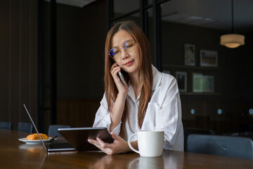 Asian woman wearing eyeglasses talking on the phone,  looking data information at tablet, using laptop computer for work in the morning on the wooden dining table, working from home concept