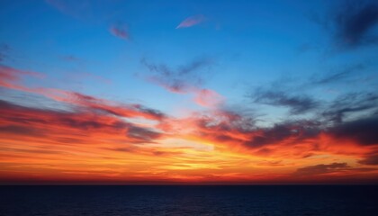 Fototapeta premium Fiery orange and blue sunset over deep blue ocean. Sun descends toward horizon line. Wispy clouds dot the sky adding depth. Calm sea reflects dramatic twilight colors.