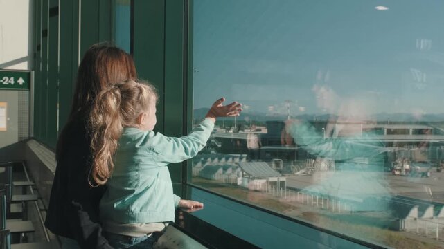 Slow motion of a mother and daughter waving goodbye at the airport window as a plane departs. Emotional farewell moment captured through glass with reflections of the runway and sky. 4k footage