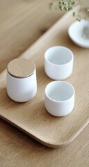 Minimalist tea set on a wooden tray. White ceramic cups and container with a wooden lid, arranged neatly. Subtle light and soft focus