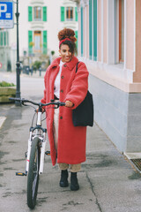 Outdoor fashion portrait of pretty african woman walking with bicycle , wearing long red coat