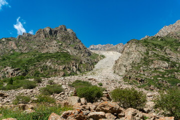 Steep rocky mountains with a prominent scree slope, scattered boulders, green shrubs, clear blue...