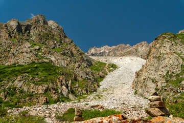 Steep mountain pass with rugged cliffs, loose scree slope, scattered boulders, lush alpine grass,...