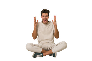 Young caucasian man sitting on the floor isolated on white background celebrating a victory or success, he is surprised and shocked.