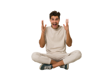 Young caucasian man sitting on the floor isolated on white background celebrating a victory or success, he is surprised and shocked.