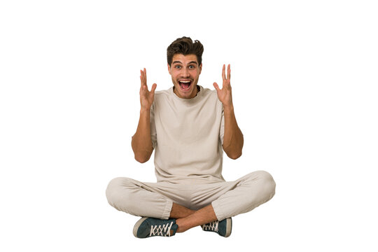 Fototapeta Young caucasian man sitting on the floor isolated on white background celebrating a victory or success, he is surprised and shocked.