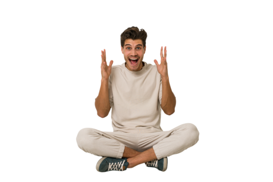 Young caucasian man sitting on the floor isolated on white background celebrating a victory or success, he is surprised and shocked.