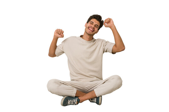 Young caucasian man sitting on the floor isolated on white background celebrating a special day, jumps and raise arms with energy.