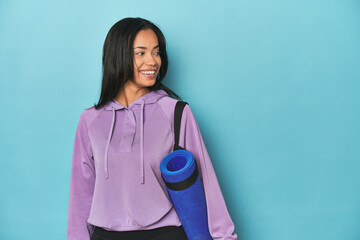 Filipina athlete with yoga mat on blue looks aside smiling, cheerful and pleasant.