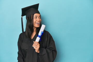 Filipina graduate with diploma on blue studio points with thumb finger away, laughing and carefree.