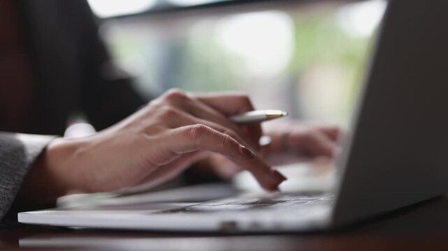 Professional businesswoman typing on laptop keyboard while holding pen, focusing on work in modern, light filled office environment with blurred background