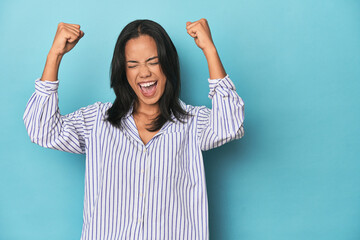 Filipina young woman on blue studio raising fist after a victory, winner concept.