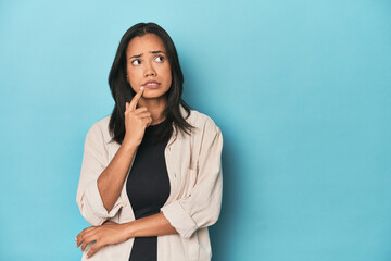 Filipina young woman on blue studio looking sideways with doubtful and skeptical expression.