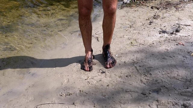 Man's feet stained with black mud stand by a river, a stark representation of pollution's effect on nature.