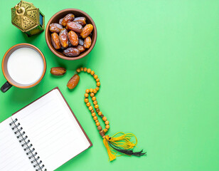 Flat lay featuring dates in a wooden bowl, milk, prayer beads with tassels, a lit lantern, and an open blank notebook on a green surface. A peaceful arrangement evoking spirituality and reflection