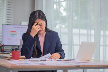 Exhausted businesswoman sitting at desk in office surrounded by documents and charts, facing...