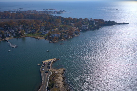 Aerial view of the sunlit water meeting the rocky coastline and autumn-tinged trees, with a pier stretching into the calm bay, Branford, Connecticut, United States.