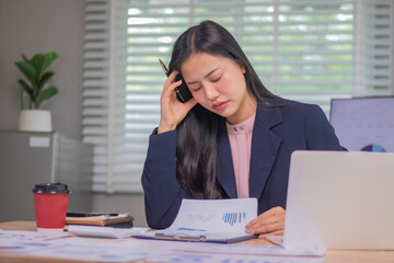 Exhausted businesswoman sitting at desk in office surrounded by documents and charts, facing...