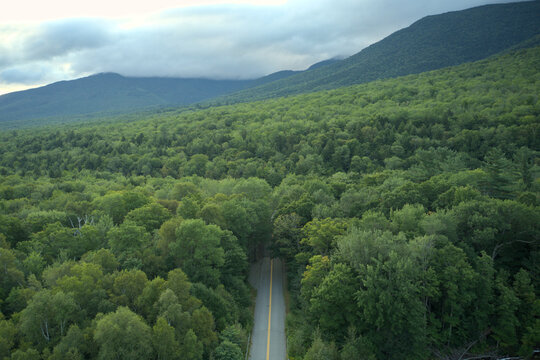 Aerial view of a road cutting through verdant forest towards the majestic mountains under a cloudy sky, Gorham, New Hampshire, USA.