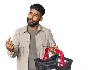 Hispanic male holding shopping basket in studio pointing with finger at you as if inviting come...